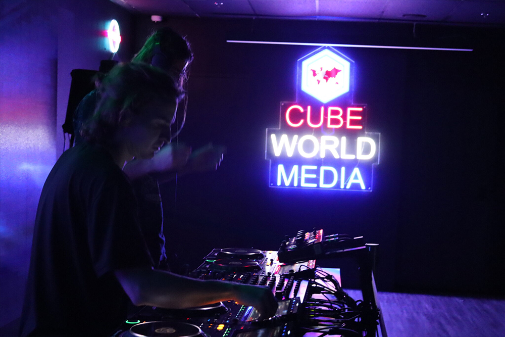 A DJ performs behind a lit controller and mixer in a dark room with a glowing neon Cube World Media sign.
