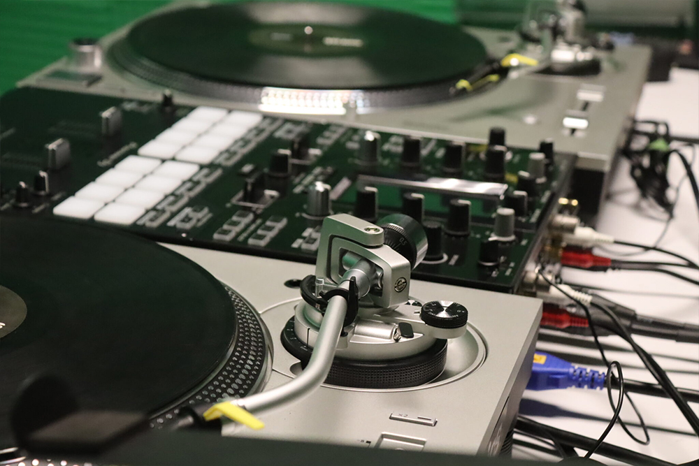 Two silver DJ turntables beside a battle mixer with white pads and knobs, with red, white, and blue cables plugged in.