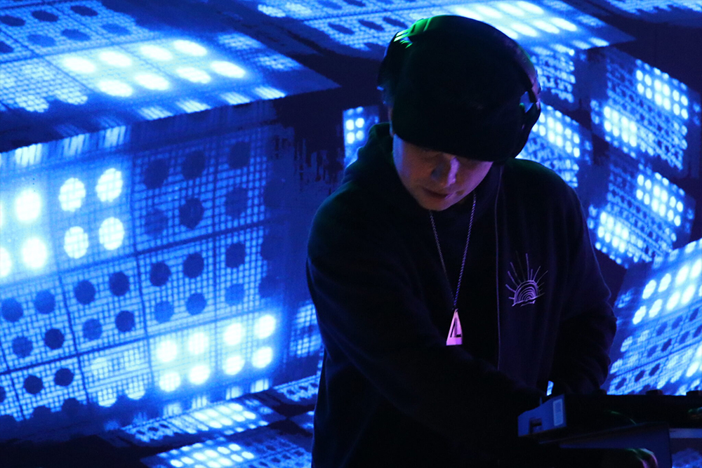 Person in beanie and headphones leaning over DJ gear surrounded by glowing blue LED panels with dot patterns
