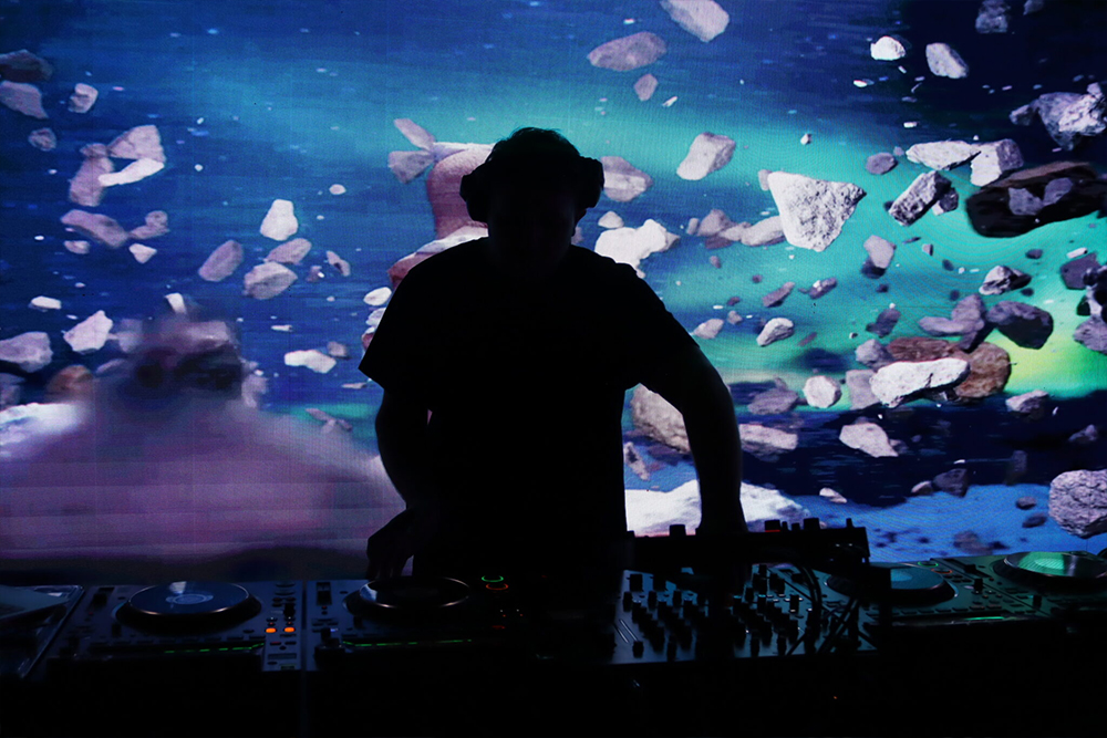 Silhouetted DJ with headphones at a two-deck controller and mixer in front of a large LED wall showing an underwater scene.