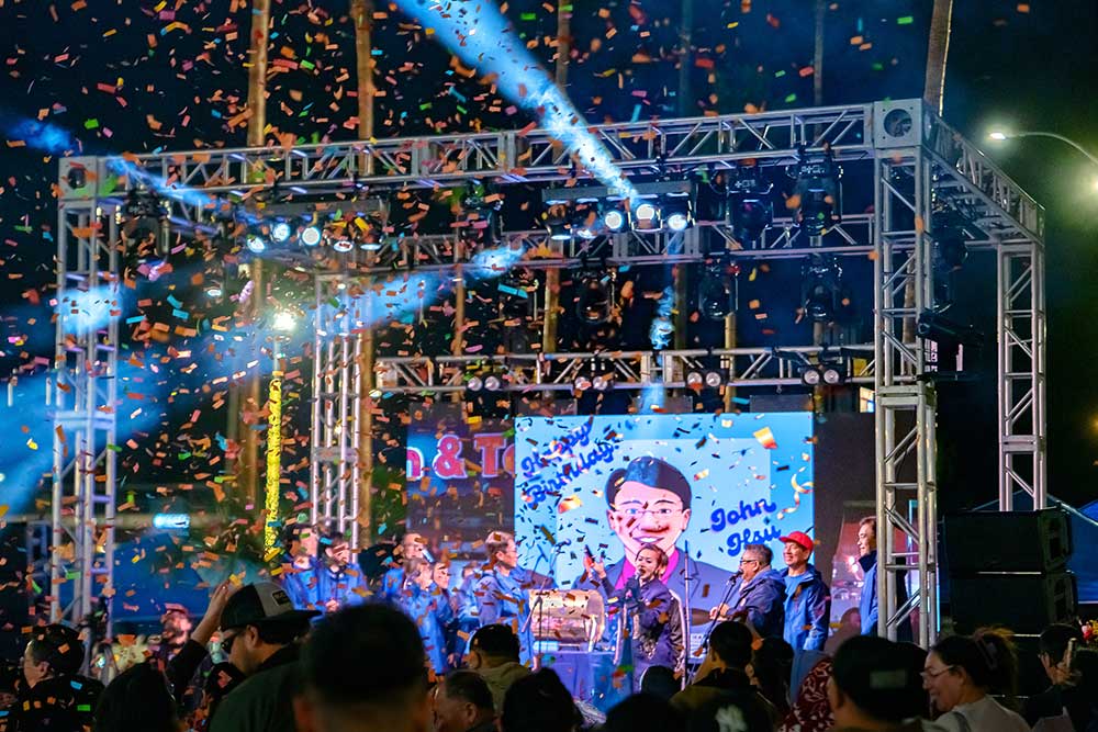 Outdoor truss stage at night with blue lights, LED screens, performers in blue jackets, and confetti over a crowd.