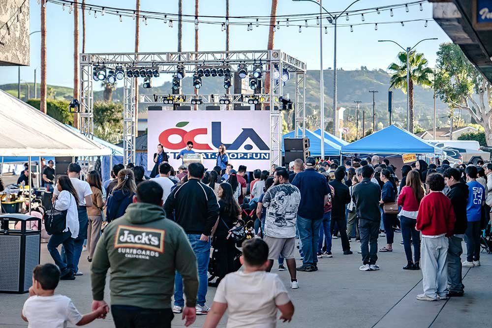 Outdoor event stage with truss lighting, an LED screen showing a logo, PA speakers, and a crowd facing it.