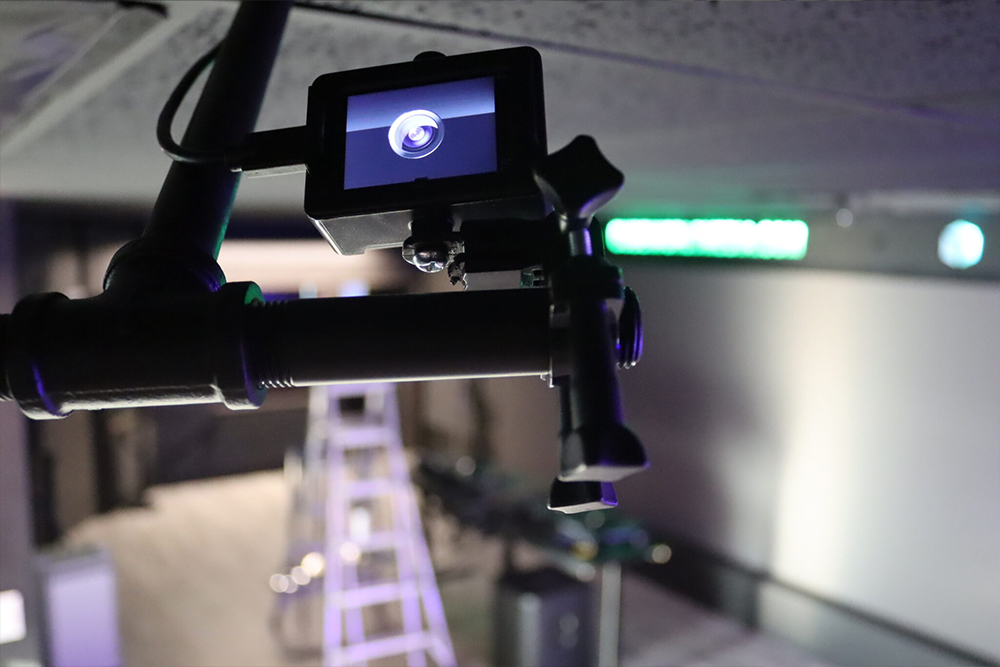 Video camera on black pipe rigging with its rear screen on, in a dark venue with a ladder and colored lighting.
