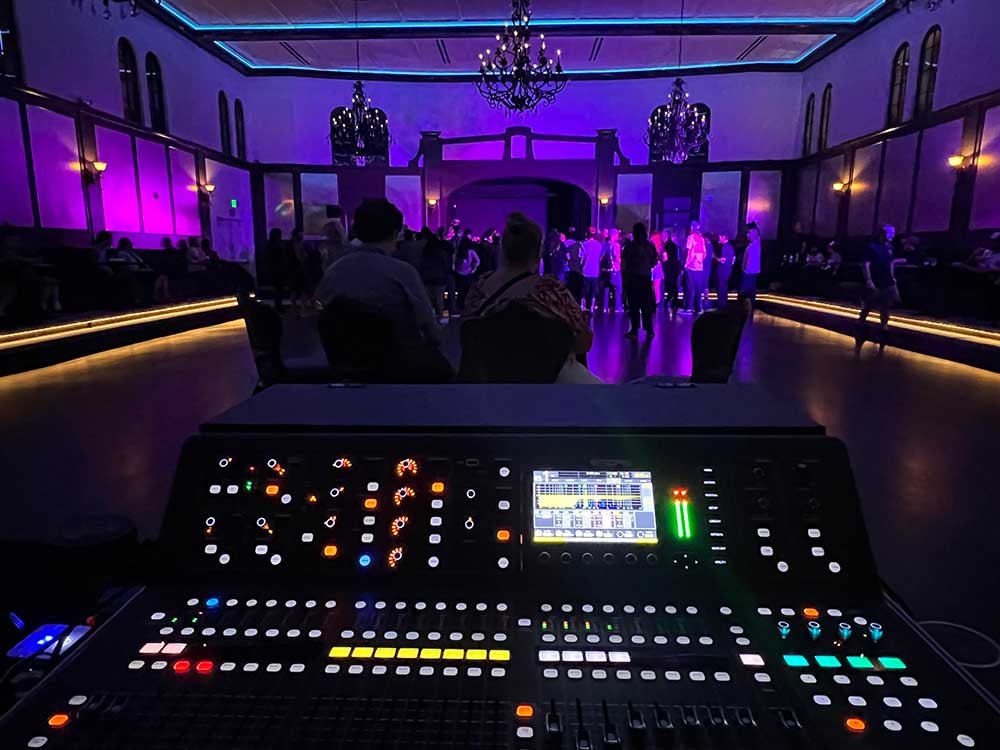 Digital audio mixing console at front of house facing a dim ballroom with chandeliers, purple uplighting, and a crowd of guests.