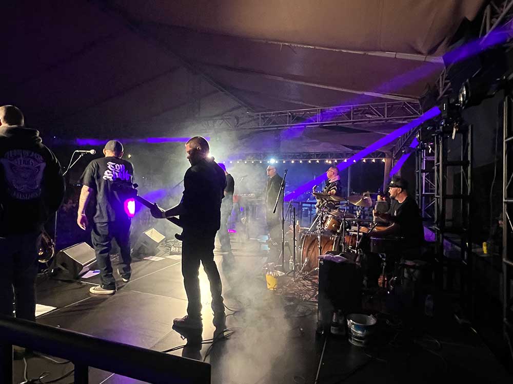 Band performing on a night stage under a tent roof with purple lighting, festoon lights, monitors, and a drum kit.