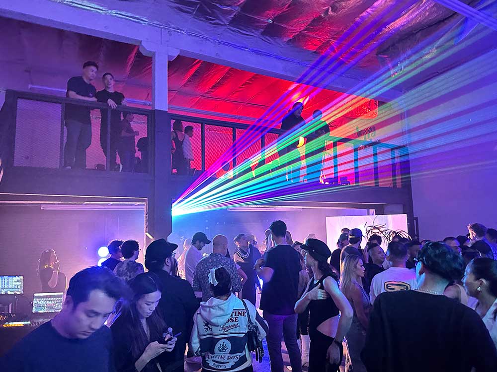 Crowded indoor venue with colorful laser beams fanning across the room and people on a mezzanine above the main floor.