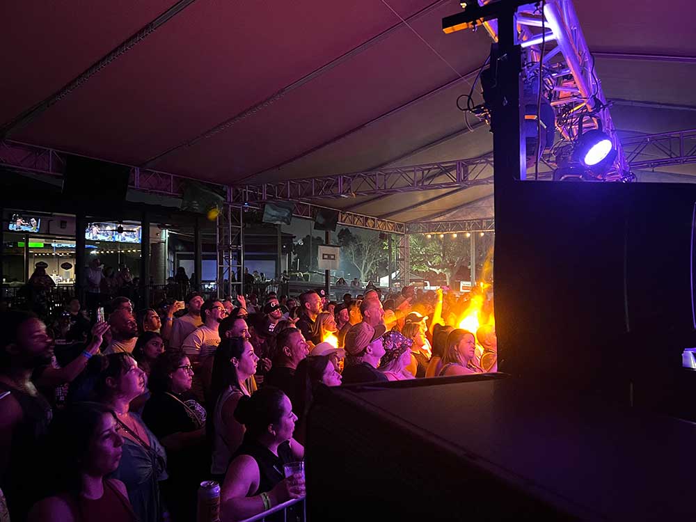 Crowd at a nighttime tented concert with purple moving-head light and speaker stack on truss, warm stage lighting.