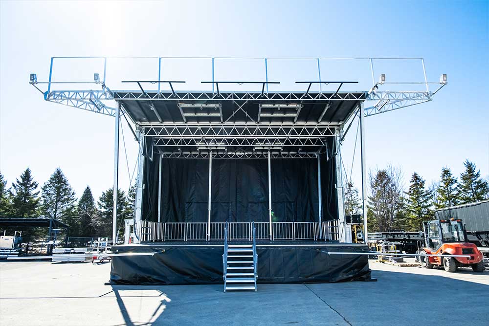 Mobile trailer stage with aluminum truss roof, black backdrop and skirting, edge railing, and front stairs on an outdoor paved lot.