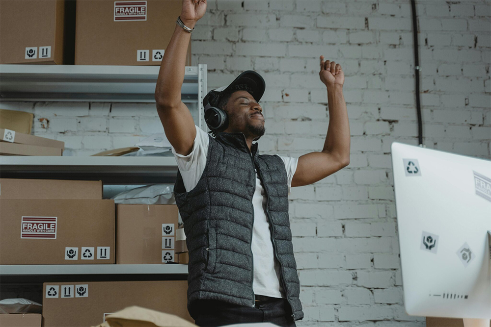 Person in a vest and headphones raises both arms in a warehouse beside shelves of boxes marked Fragile.
