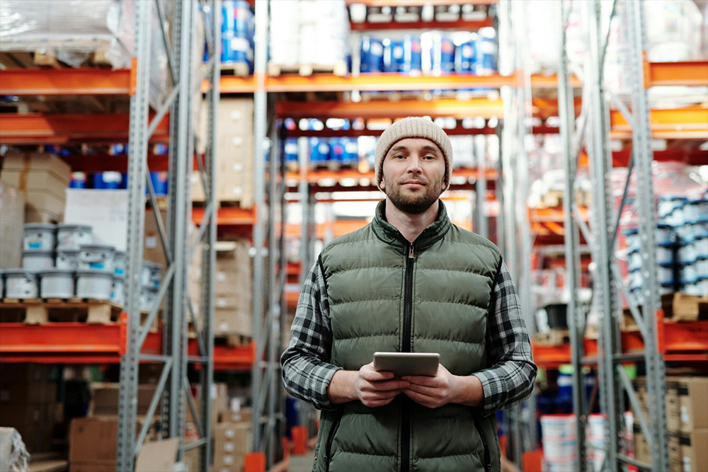 Person in a beanie and green vest holds a tablet in a warehouse aisle between tall pallet racks stacked with boxes and drums.